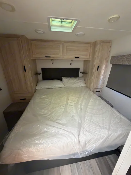 RV rental bedroom: New bed (in plastic), dark headboard, wood cabinets, skylight, and reading lights for comfortable travel.. Forest River Sunseeker LE 2024