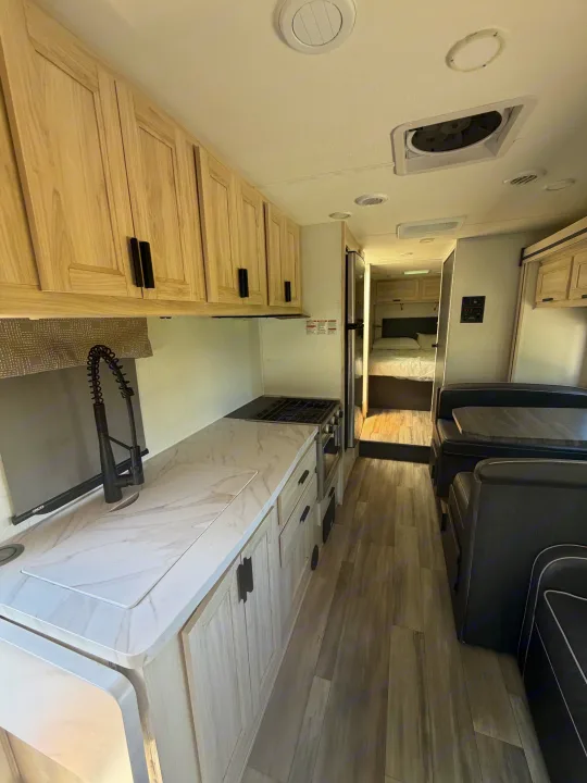 Inside a modern RV rental, featuring a full kitchen with light wood cabinets, a dining booth, and a peek into the bedroom.. Forest River Sunseeker LE 2024