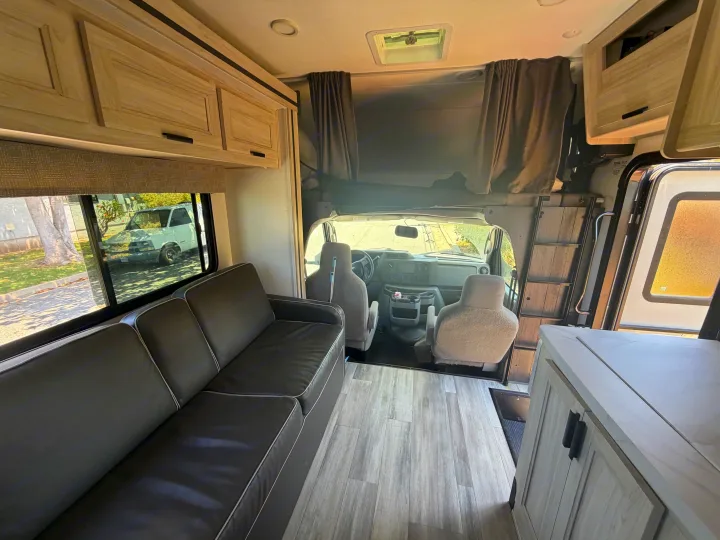 Inside this RV rental: modern living space with dark couch, wood-look floor, kitchenette, and overhead bunk. Driver's cockpit visible.. Forest River Sunseeker LE 2024