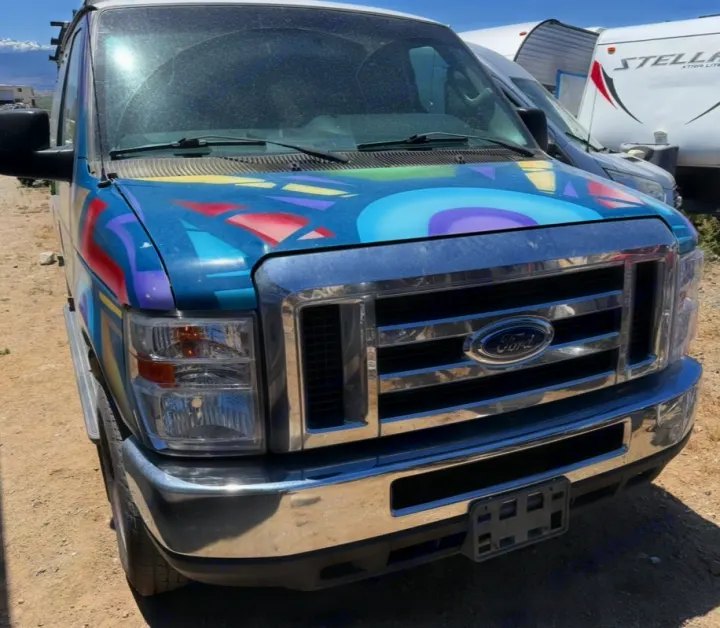 A blue Ford van with custom colorful abstract paint on its hood, a dusty windshield, and a chrome grille. Appears to be an RV rental.. Ford E350 Box Van 2013