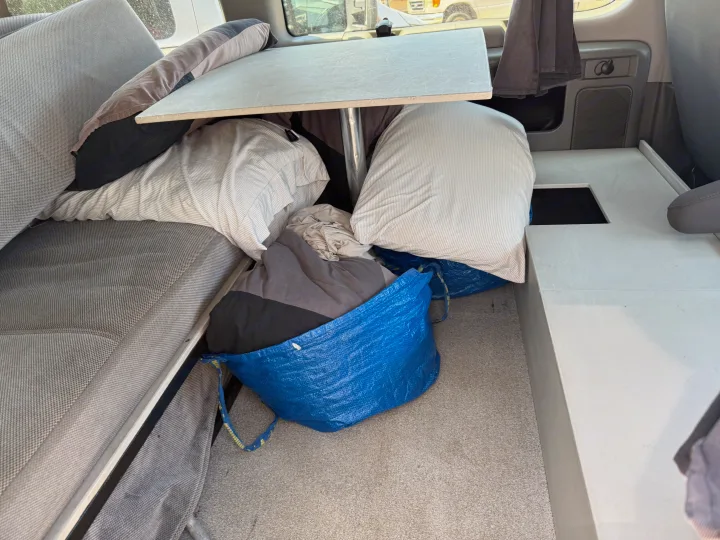 Inside this rv rental, a compact living area features a bed, dining table, and personal belongings stored in blue bags.. Ford E350 Box Van 2013