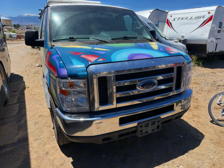 A custom-painted blue Ford van, possibly an RV rental, sits in a dusty lot with snowy mountains and other trailers.. Ford E350 Box Van 2013