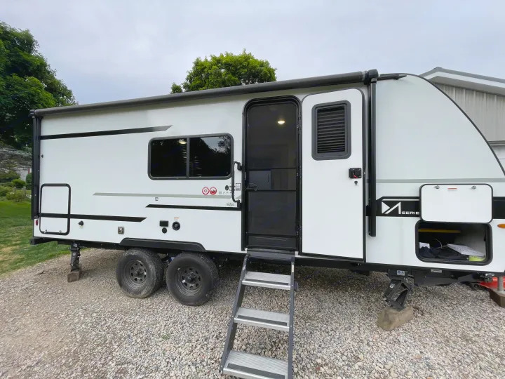 White travel trailer RV rental parked on gravel with open door, steps, awning, and front storage compartment.. Winnebago M Series 2025