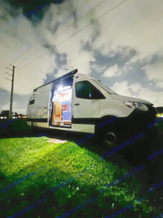 Night scene: a white rv rental camper van on grass, side door open to reveal a brightly lit, cozy interior.. Mercedes-Benz Sprinter 2023