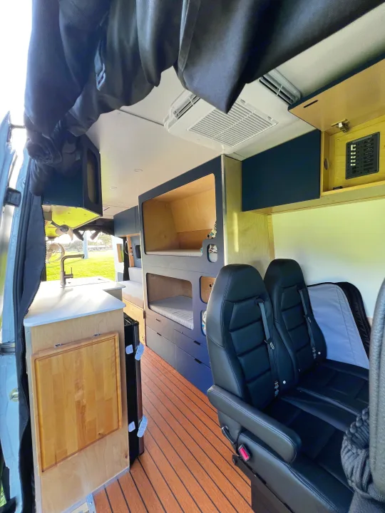 Interior of a custom rv rental van with a kitchen, bunk beds, seating, wood-slat flooring, and an overhead AC unit.. Mercedes-Benz Sprinter 2023