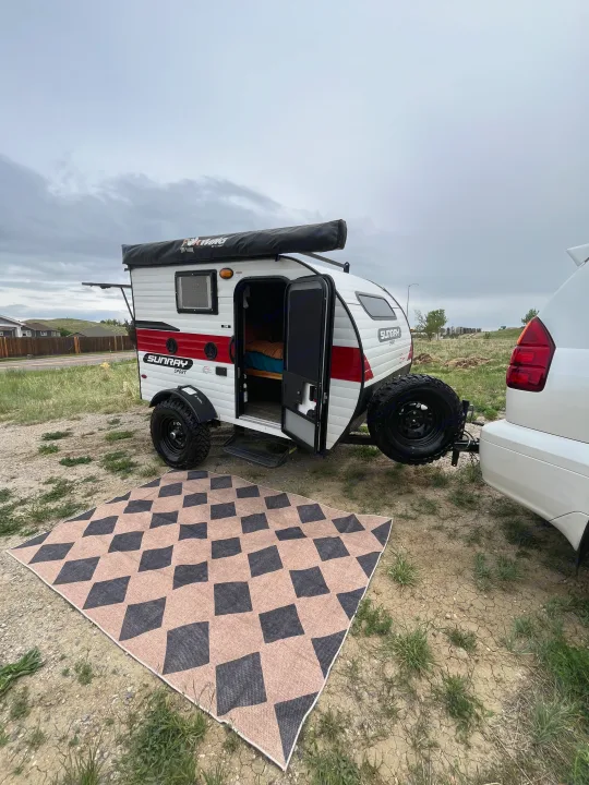 An RV rental: A small white & red teardrop camper hitched to an SUV, door open to a bed, next to an outdoor mat.. Sunset Park RV SunRay 2021