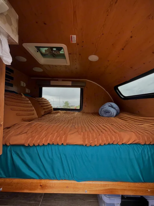 Cozy rv rental interior with warm wood panels. A bed with brown textured bedding, teal sheets, and a blue blanket is set for comfort.. Sunset Park RV SunRay 2021
