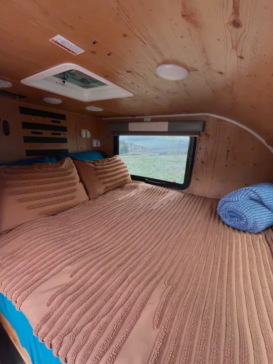 Cozy RV rental sleeping area with wood-paneled walls. Features a textured bed, pillows, and a window view of green fields.. Sunset Park RV SunRay 2021