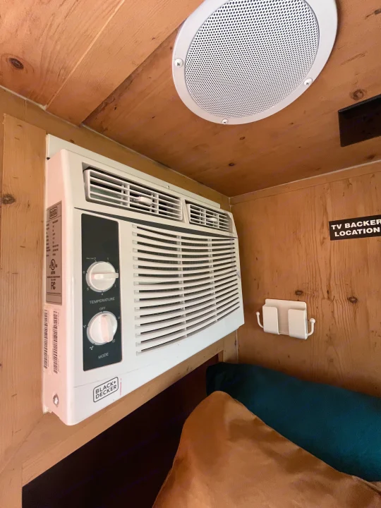 RV rental cabin: Wall-mounted Black+Decker AC unit, ceiling speaker, and wooden interior details, including a "TV BACKER LOCATION" sign.. Sunset Park RV SunRay 2021