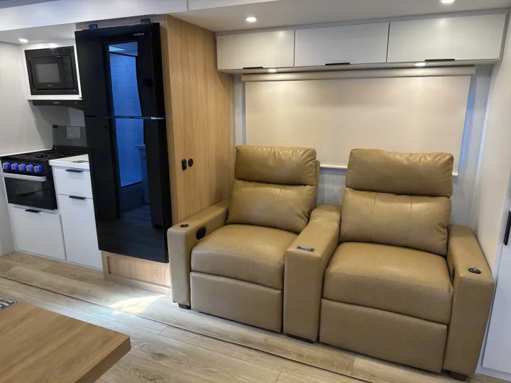 This modern rv rental interior features a sleek kitchenette, black refrigerator, light wood accents, and two comfy tan recliners.. Winnebago Thrive 2026