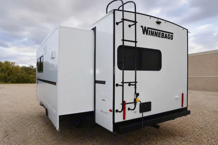 Rear view of a white Winnebago RV rental with an extended slide-out, a black ladder, and a power cord plugged in, parked on gravel.. Winnebago Thrive 2026