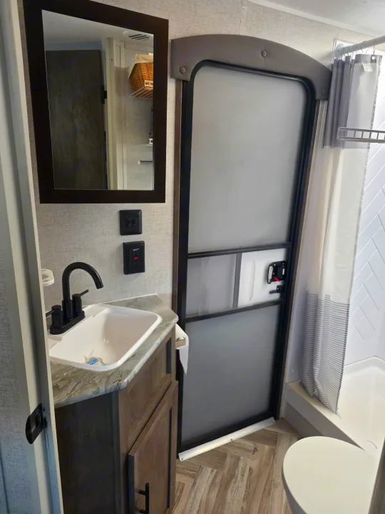 RV rental bathroom features a compact dark wood vanity, white sink, black faucet, mirror, shower, and toilet for convenience.. Forest River Wildwood 2021