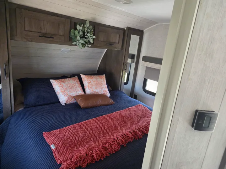 Cozy RV rental bedroom with navy bedding, decorative pillows, and a coral throw, featuring rustic cabinetry and windows with shades.. Forest River Wildwood 2021