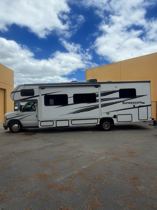 White Class C RV rental, a Sunseeker motorhome, parked on asphalt with tan buildings & blue sky with clouds.. Forest River Sunseeker LE 2024