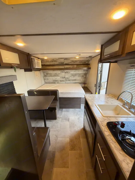 This rv rental interior features a cozy rear bed, dinette booth, and kitchen with marble-look counter, sink, and stovetop.. Coleman 17B Dutchmen 2022