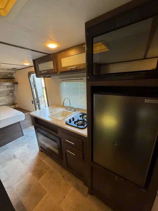 This RV rental's compact kitchen includes a sink, two-burner stove, microwave, and fridge, with a bed visible. Note wear on surfaces.. Coleman 17B Dutchmen 2022