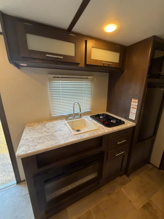 Explore this RV rental's compact kitchen featuring a sink, stovetop, cabinets, refrigerator, and an electric fireplace.. Coleman 17B Dutchmen 2022