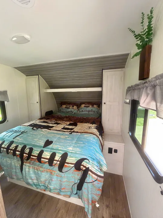 Cozy RV rental bedroom with "Happy Camping" bedding, built-in storage, corrugated headboard, and windows.. Heartland RVs Trail Runner 2024