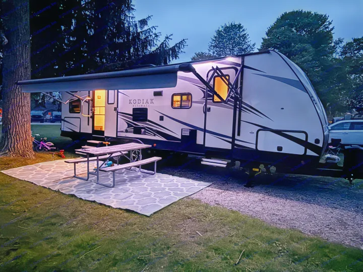 Cozy rv rental at night, with warm interior lights, an awning, picnic table, and outdoor rug set for camping.. Kodiak Ultimate 288BHSL 2018