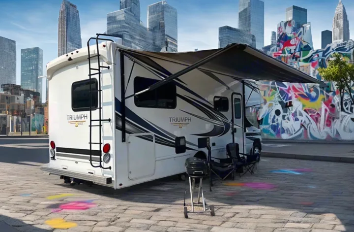 A white "Triumph" RV rental with an extended awning, chairs, and a grill, parked in an urban setting with graffiti and city buildings.. Nexus Triumph 23T 2020