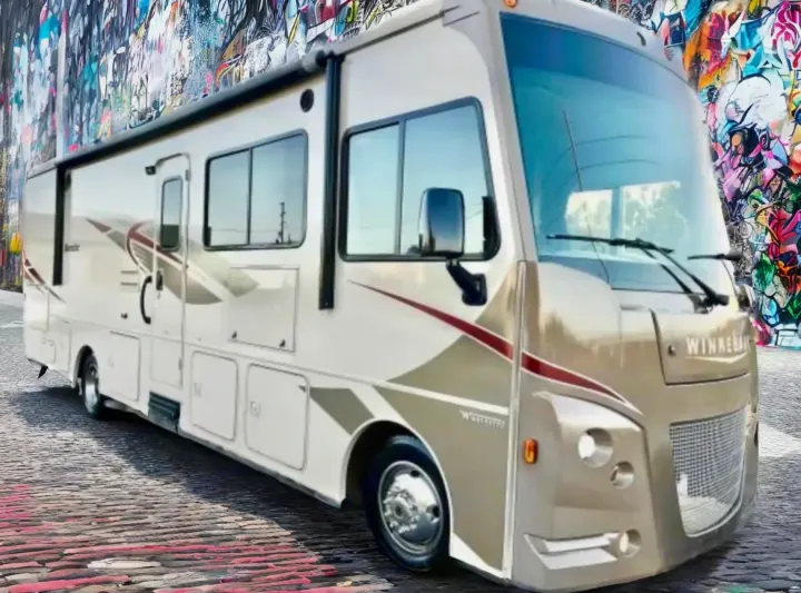 A beige Winnebago motorhome, ideal for rv rental, is parked on a brick street against a vivid graffiti wall backdrop.. Winnebago Sunstar 2016