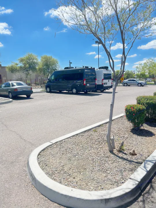 A sunny parking lot features a dark Jayco Swift and a white Solis camper van, indicating RV rental services, alongside other vehicles.. Jayco Swift Li 2023