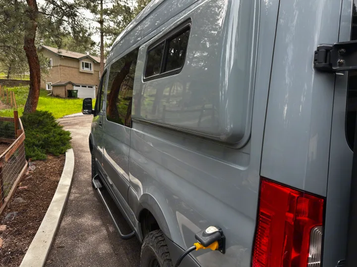 Grey RV rental van, seen from the side, charging via shore power. Parked on a driveway near a house and trees.. Mercedes-Benz Sprinter 2020