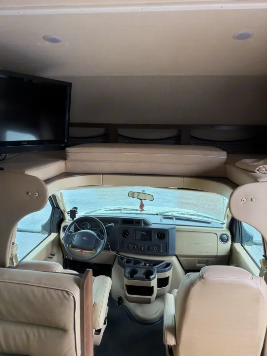 Inside an rv rental: view from living area to the front cockpit with Ford dash, TV, and overhead bunk. Beige interior.. Jayco Redhawk 2020