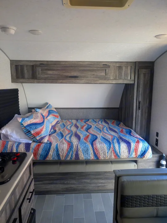 Relax in this cozy RV rental's bedroom, featuring a vibrant patterned quilt, practical overhead cabinets, and a nearby kitchen view.. Forest River Wildwood FSX Plantium 2022