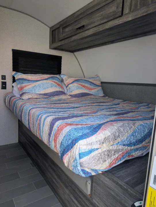 Cozy RV rental bedroom featuring a bed with a colorful wavy quilt, pillows, overhead storage, window blind, and dark tile floor.. Forest River Wildwood FSX Plantium 2022