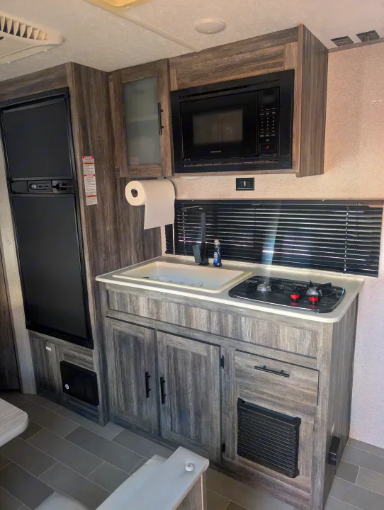 Compact rv rental kitchen featuring a fridge, microwave, sink, two-burner stove, and storage cabinets.. Forest River Wildwood FSX Plantium 2022