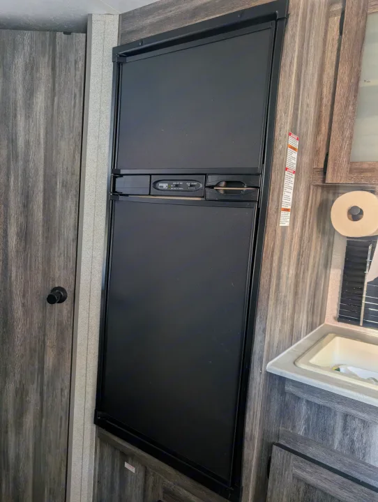 RV rental interior with a tall black refrigerator, wood-grain cabinets, door, and small sink area.. Forest River Wildwood FSX Plantium 2022
