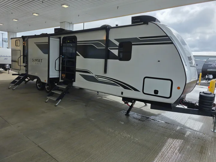 A modern white Sunset Trail RV, featuring steps and graphics, is parked under a canopy. Ideal for your next rv rental adventure.. Crossroads RV Sunset Trail Super Lite 2025