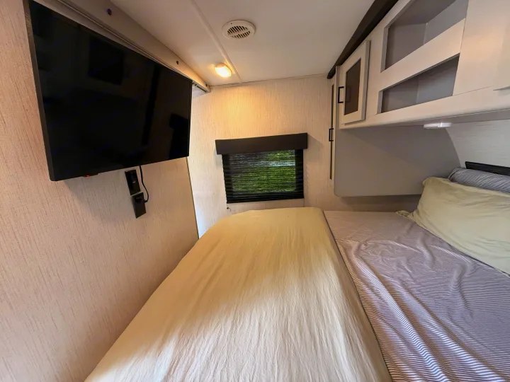 RV rental bedroom. A wall-mounted TV, window with blinds, and overhead cabinets are seen above a bed with striped bedding.. Heartland RVs North Trail 2022