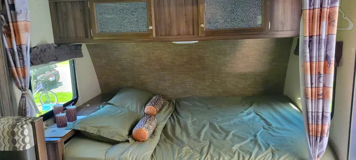Inside an RV rental: a bed with green sheets, log pillows, overhead cabinets, and a window looking out at an ATV.. Coachmen Apex Nano 2017