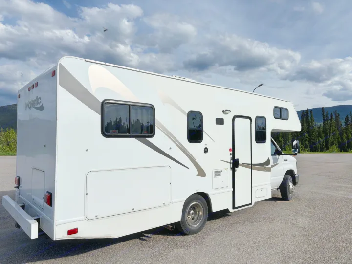 This rv rental, a white Majestic Class C, features graphic decals & is parked on asphalt with a cloudy sky.. Thor Majestic 2020