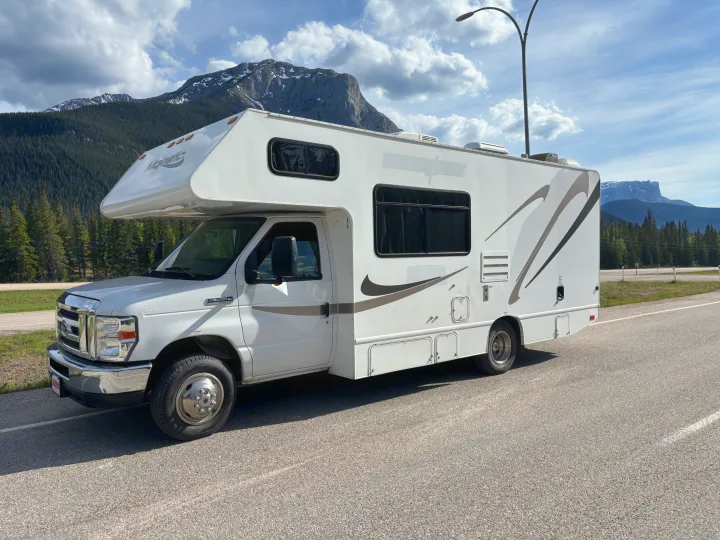 White rv rental motorhome parked on an asphalt road. Rugged mountains, pine trees, and a cloudy sky provide a scenic backdrop.. Thor Majestic 2020