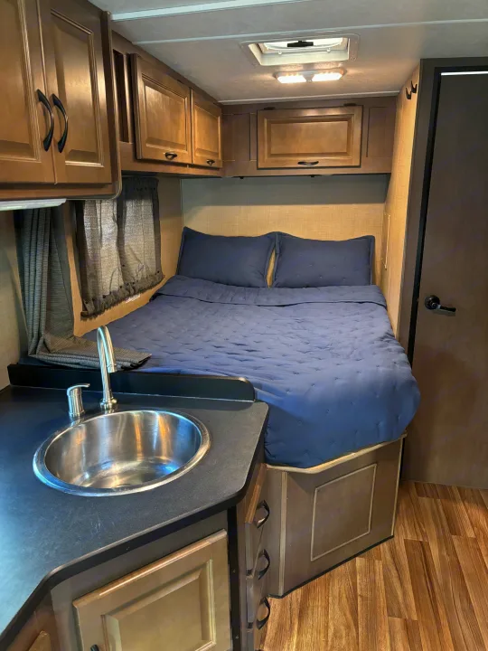 View inside a functional RV rental, featuring a compact bedroom with blue bedding, a small sink, and storage cabinets.. Thor Majestic 2020
