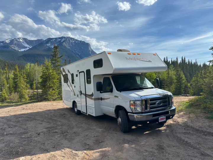 White Majestic rv rental parked on dirt with evergreen trees & stunning snow-capped mountains under a cloudy sky.. Thor Majestic 2020