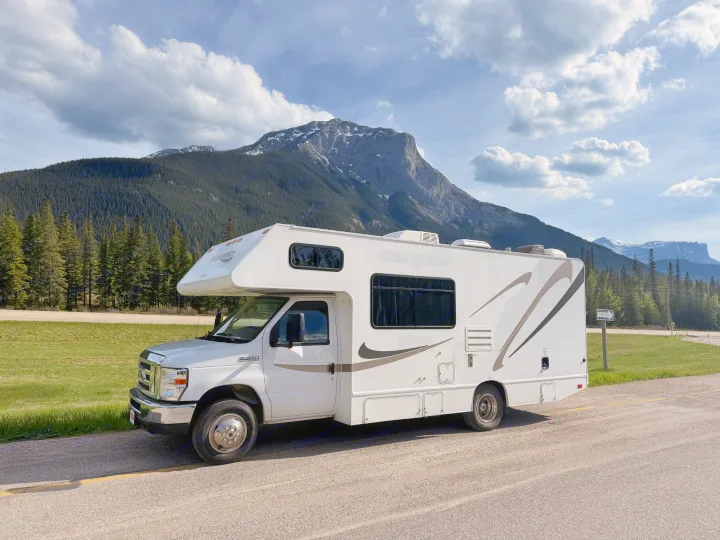 An RV rental is parked on a scenic road with towering mountains and a lush forest under a cloudy sky. Perfect for road trips.. Thor Majestic 2020