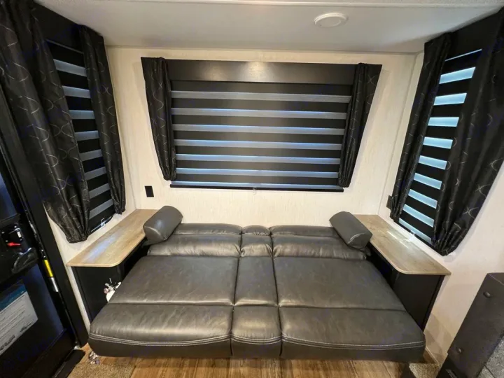 RV rental interior features a dark gray convertible couch/bed, flanked by windows with horizontal blinds and black curtains.. Forest River Cherokee Grey Wolf 2022