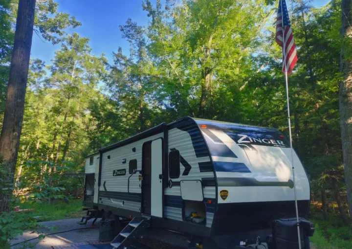 An RV rental - a Zinger travel trailer - parked in a beautiful wooded campsite with an American flag.. Crossroads RV Zinger 2021