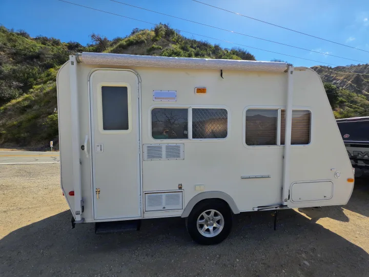 Side view of a white, compact RV rental travel trailer. It's parked on a sunny dirt road with green hills and blue sky in the background.. R-Vision Trail-Lite Bantam 2002