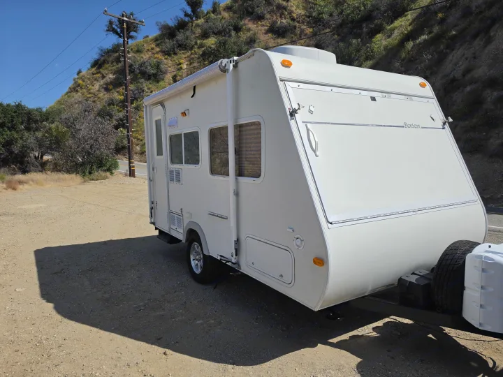 A white travel trailer, ideal for an rv rental, is parked on a sunny dirt shoulder with a green hillside in the background.. R-Vision Trail-Lite Bantam 2002