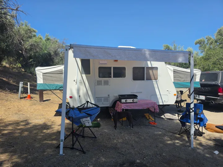 Sunny day at the campsite with an RV rental, featuring a dog, pop-up trailer, awning, and outdoor cooking setup.. R-Vision Trail-Lite Bantam 2002