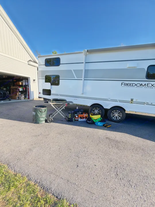 An rv rental, a Freedom EX travel trailer, is parked by a garage with a portable grill, sewer kit, and labeled water bags.. Coachmen 29 SE 2025