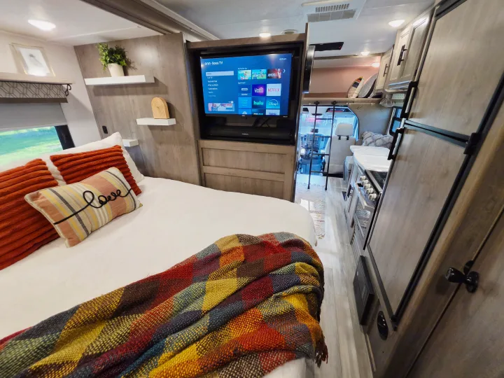 Cozy rv rental bedroom features a smart Roku TV, colorful bedding, and a glimpse into the kitchen and driver's cab.. Forest River Coachmen Prism 2018