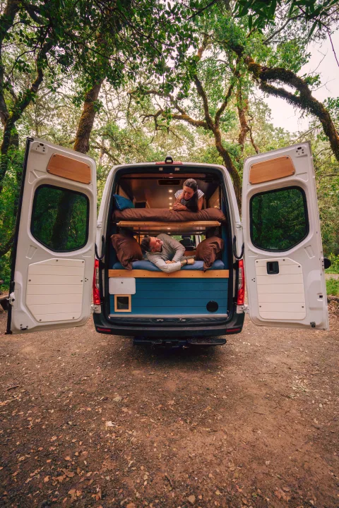 A couple relaxes in their RV rental camper van with bunk beds, rear doors open to a lush forest. Perfect for adventure.. Mercedes-Benz Sprinter 2024