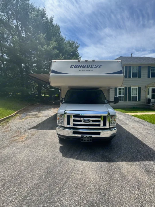 A white Ford Conquest Class C RV, likely an rv rental, parked on a sunny driveway in front of a house with trees.. Gulf Stream Conquest 2019