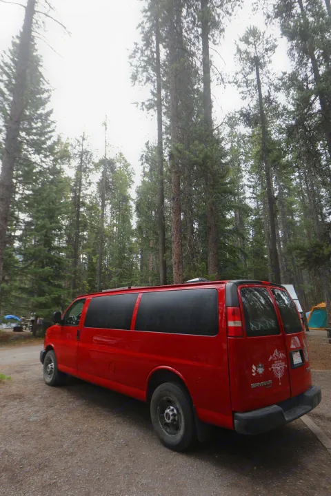 A bright red GMC Savana campervan, an ideal RV rental, parked on a gravel path in a forest campsite under towering pine trees.. Glacier Campers Vanlife Explorer 2013